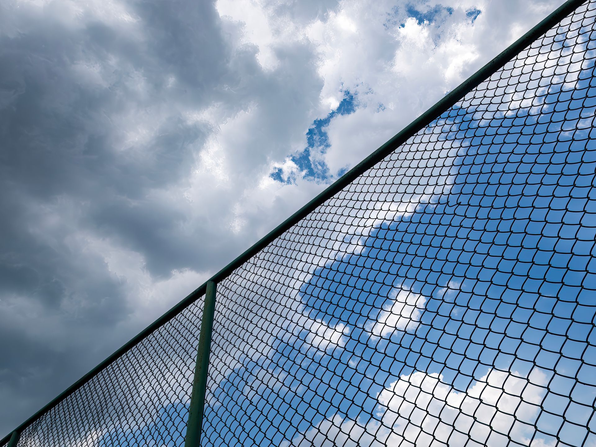 Chain-link fence against a partly cloudy sky; blue sky peeks through the fence.