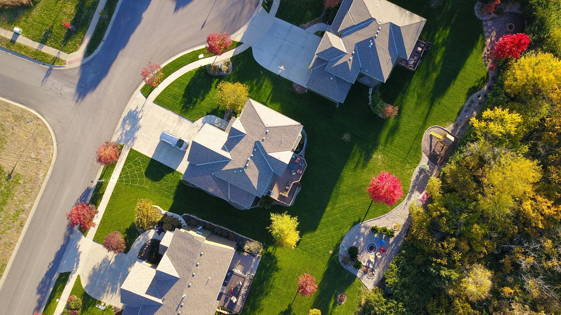 Aerial view of houses with gray roofs, green lawns, and trees with red and yellow foliage.