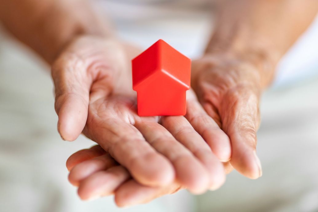 Hands cupping a small, red house model, symbolizing housing or care.
