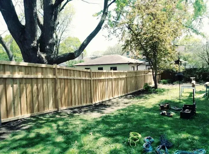 Wooden fence in a backyard with a large tree. Tools and grass visible.