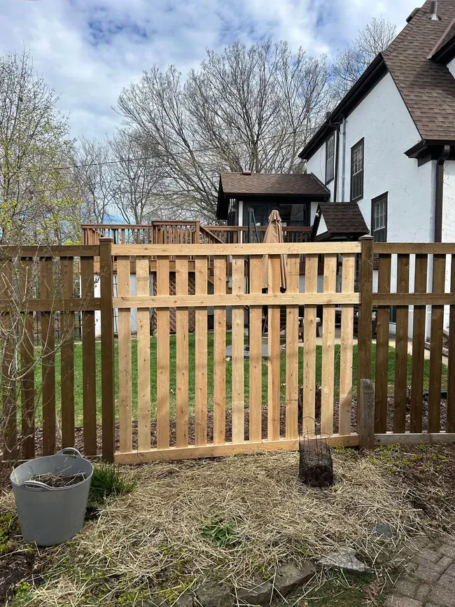 Wooden fence with gate in a yard. Tall, brown fence with a lighter colored gate in front of a white house.