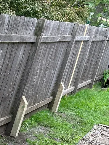 Leaning wooden fence with support beams; grassy yard, grey wood.