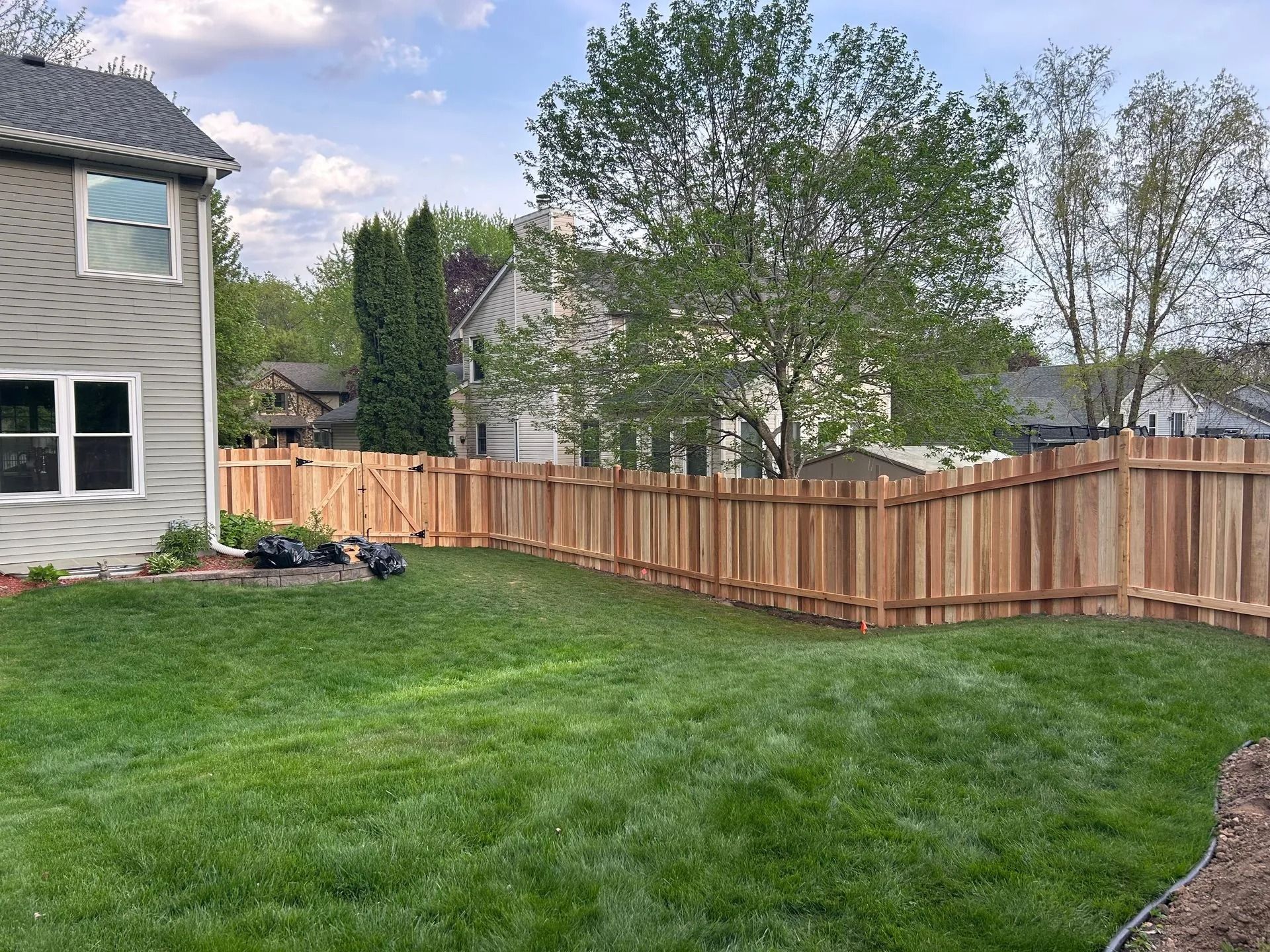 Wooden fence surrounds a grassy backyard, adjacent to a house. Cloudy sky overhead.