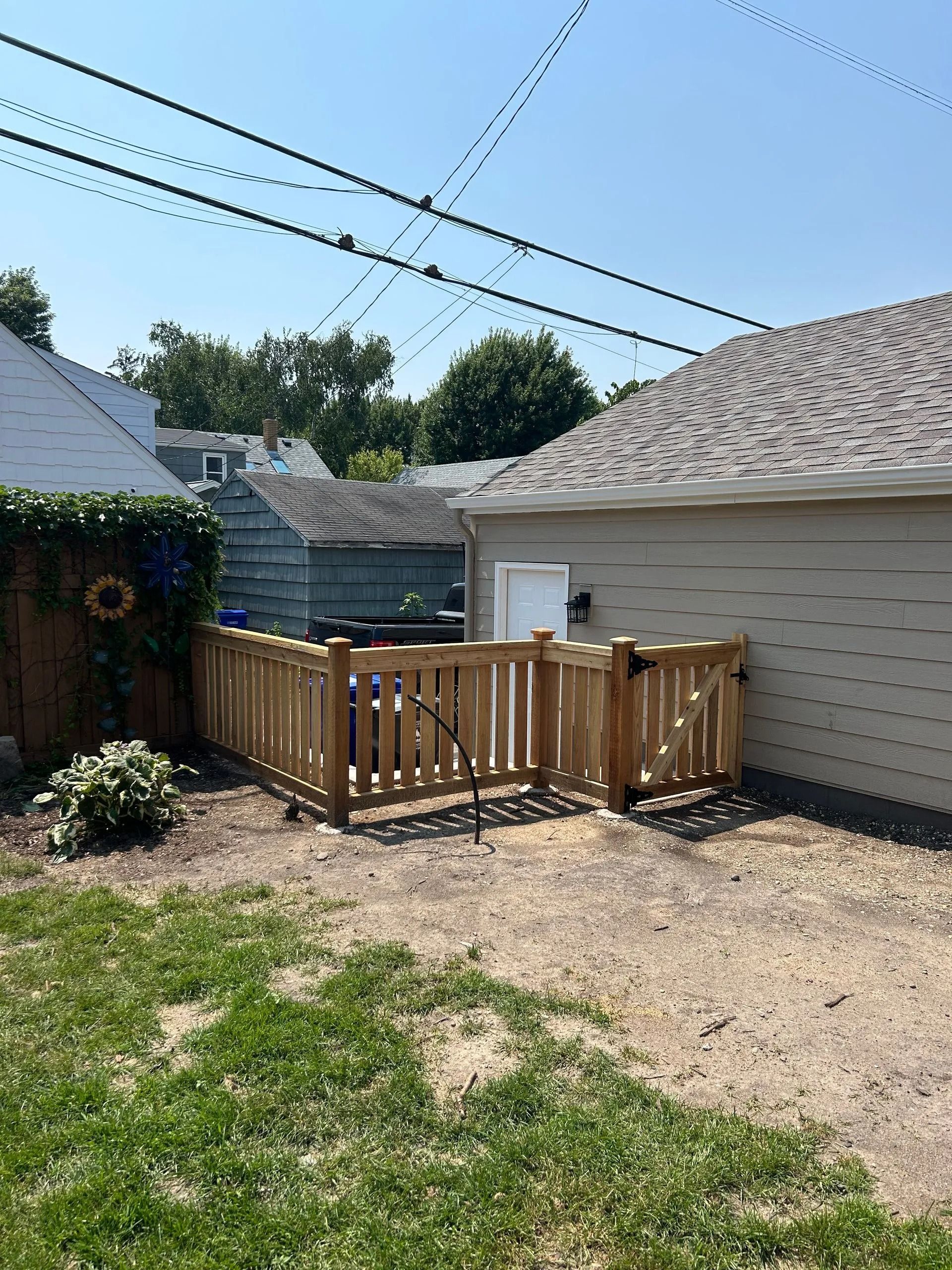Wooden fence encloses a small yard area next to a garage, with a glimpse of houses and greenery in the background.
