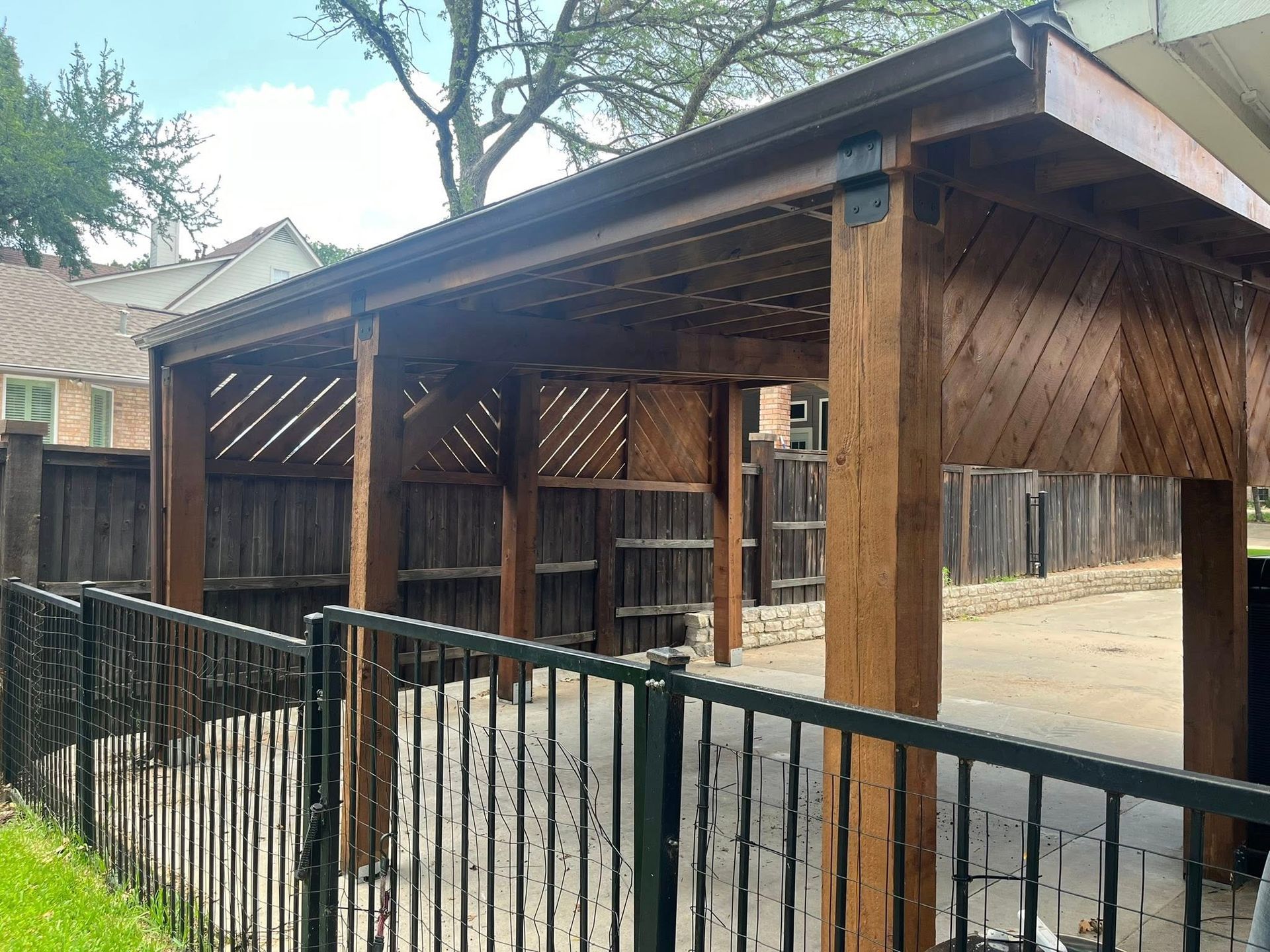 Brown wooden pergola with latticework, next to a black fence and driveway.