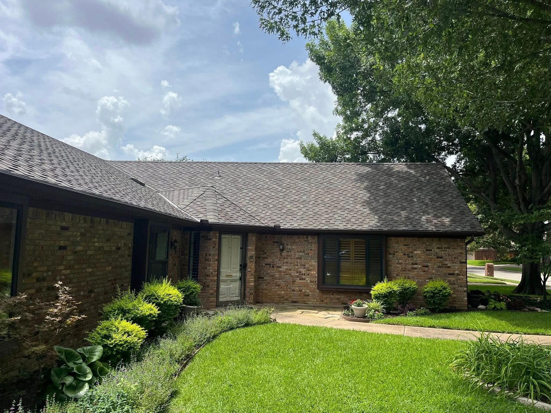 Brick house with brown roof, green lawn, and shrubs under a cloudy sky.