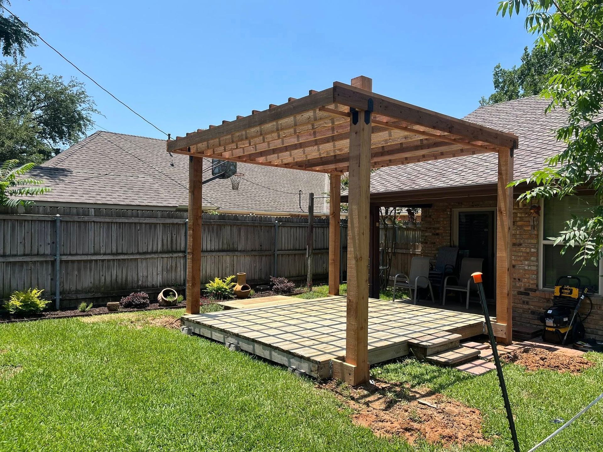 Wooden pergola over a brick patio in a backyard, next to a house with a fence.