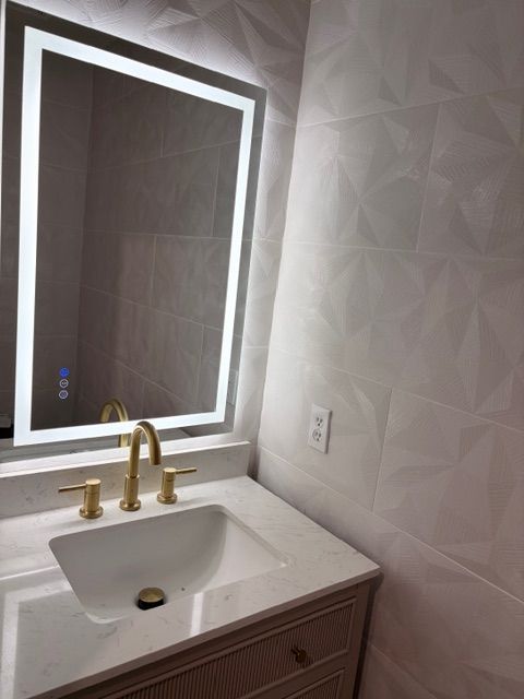 Bathroom with lighted mirror, gold faucet, white sink, and patterned wall tiles.