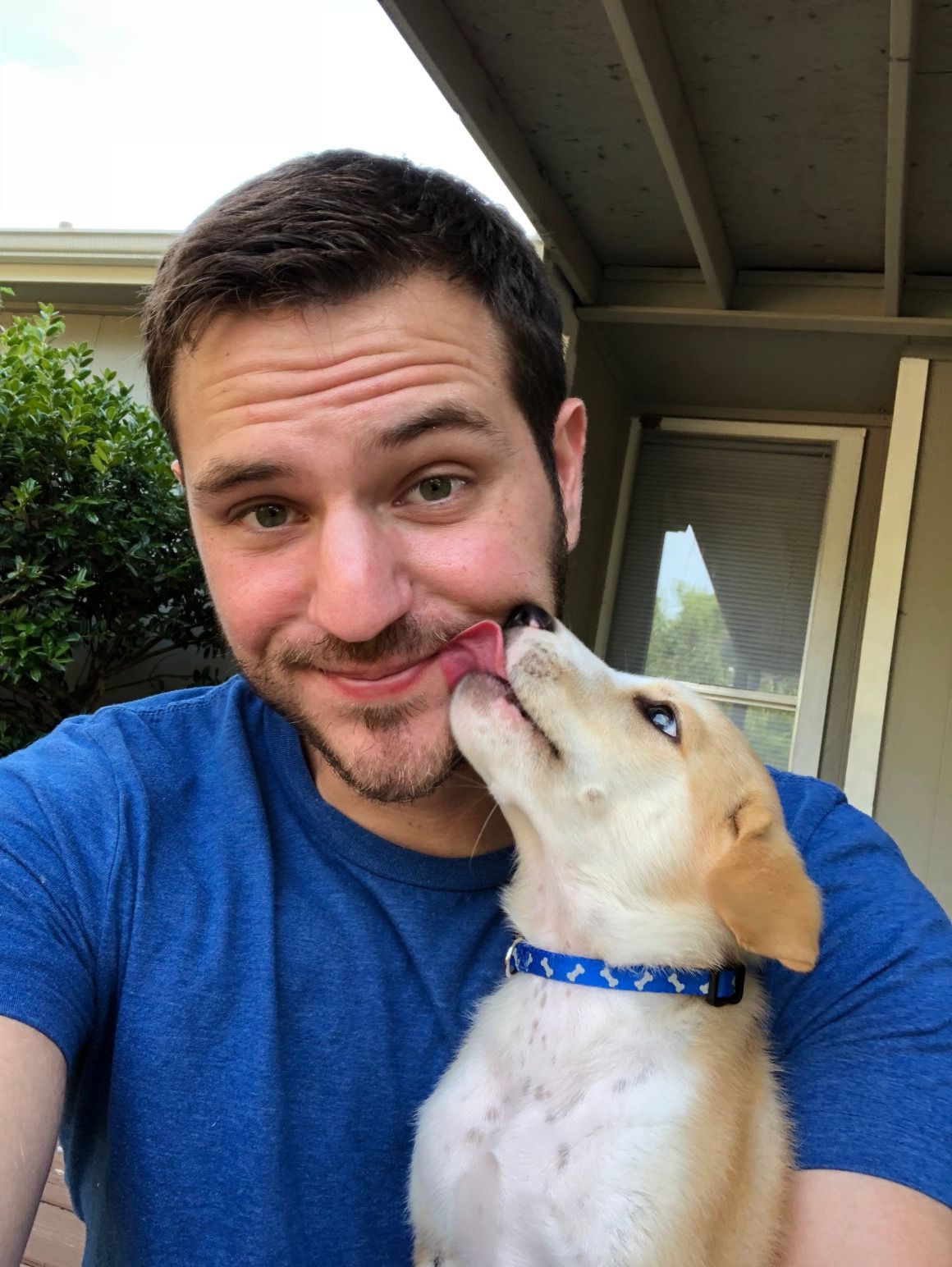 Man smiles as puppy licks his face. They are outdoors; the man wears blue.