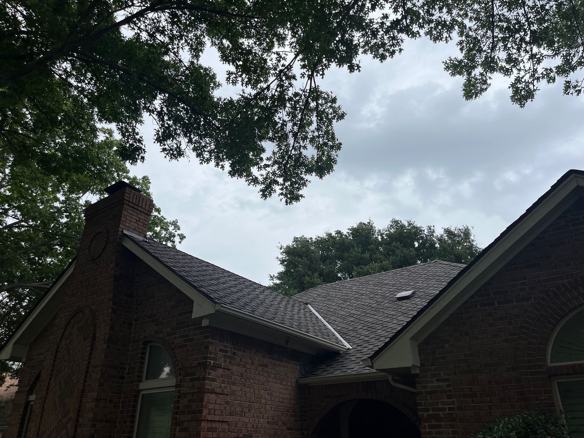 Brick house with a dark shingled roof and a chimney, under a cloudy sky.