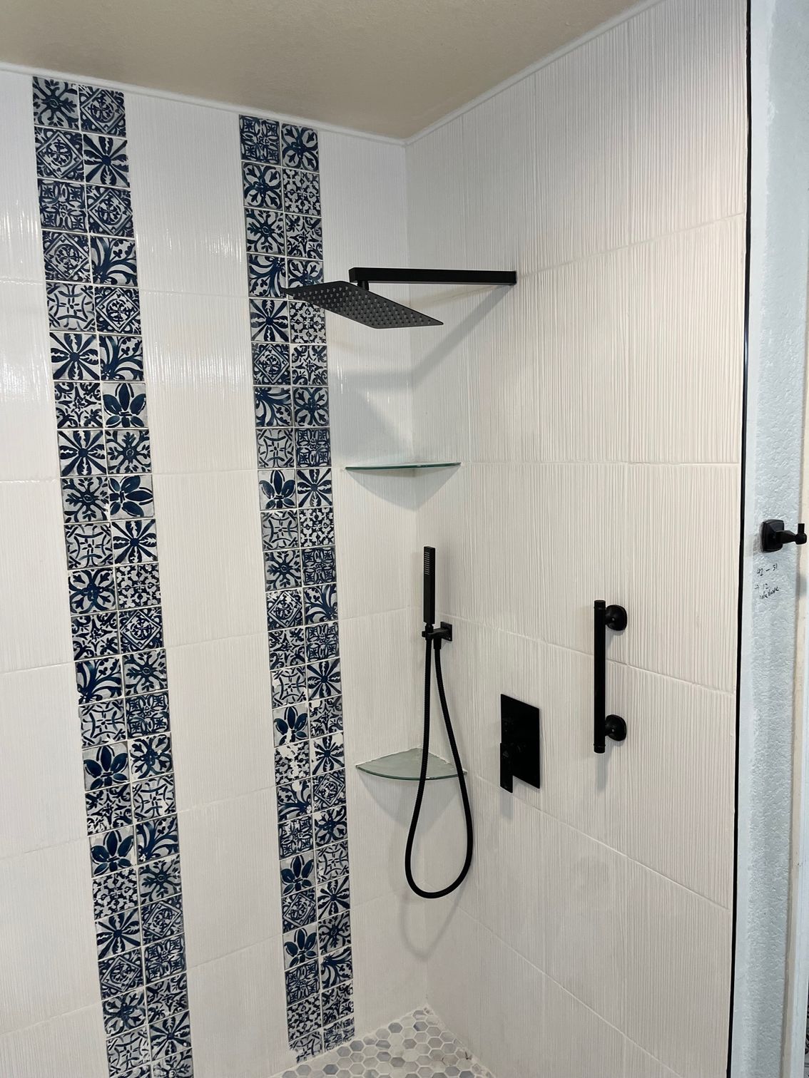 White shower with blue patterned tile accents, black fixtures, and glass shelves.