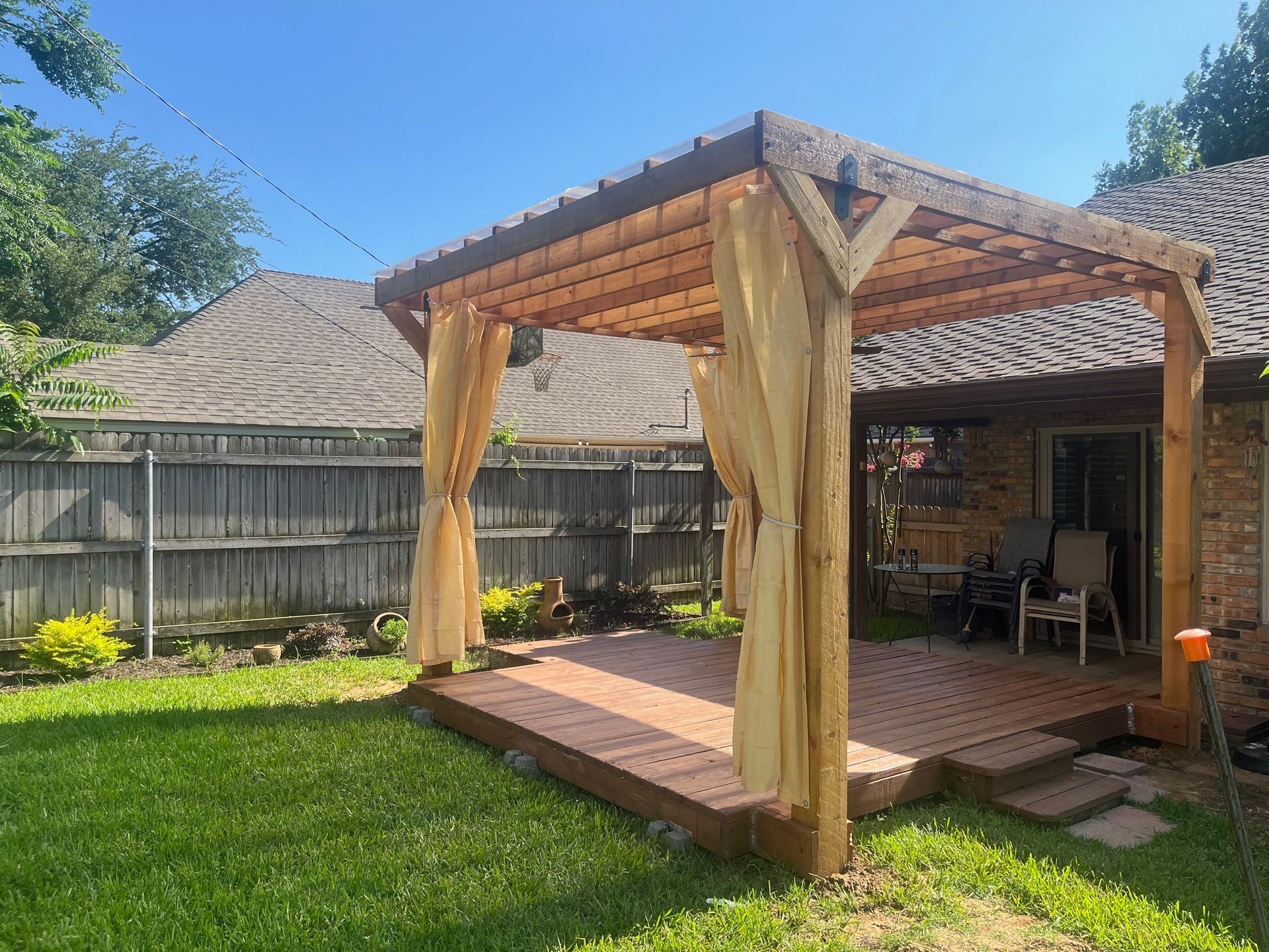 Wooden pergola on a deck with tan curtains, in a backyard, on a sunny day.