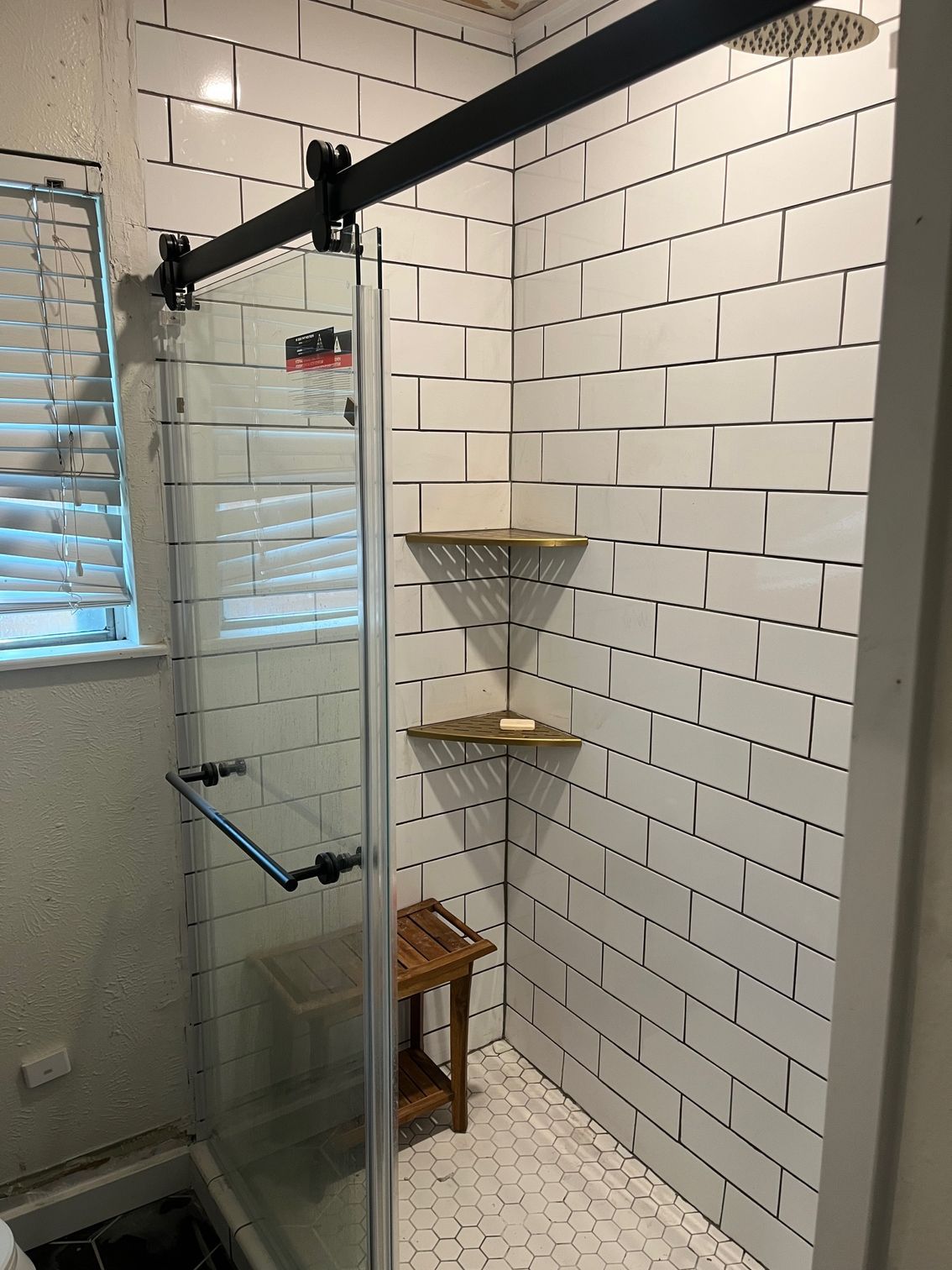 Shower stall with white brick-like tiles, glass door, wooden shelves, and a small stool.
