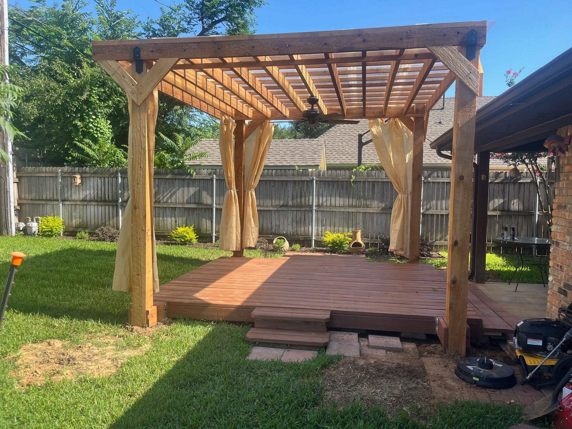 Wooden pergola with deck and curtains in a backyard.