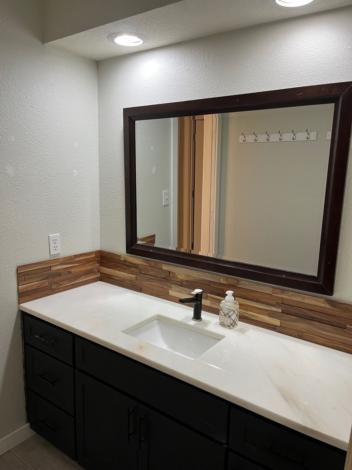 Bathroom with dark cabinets, white countertop, wooden backsplash and mirror, and black faucet.