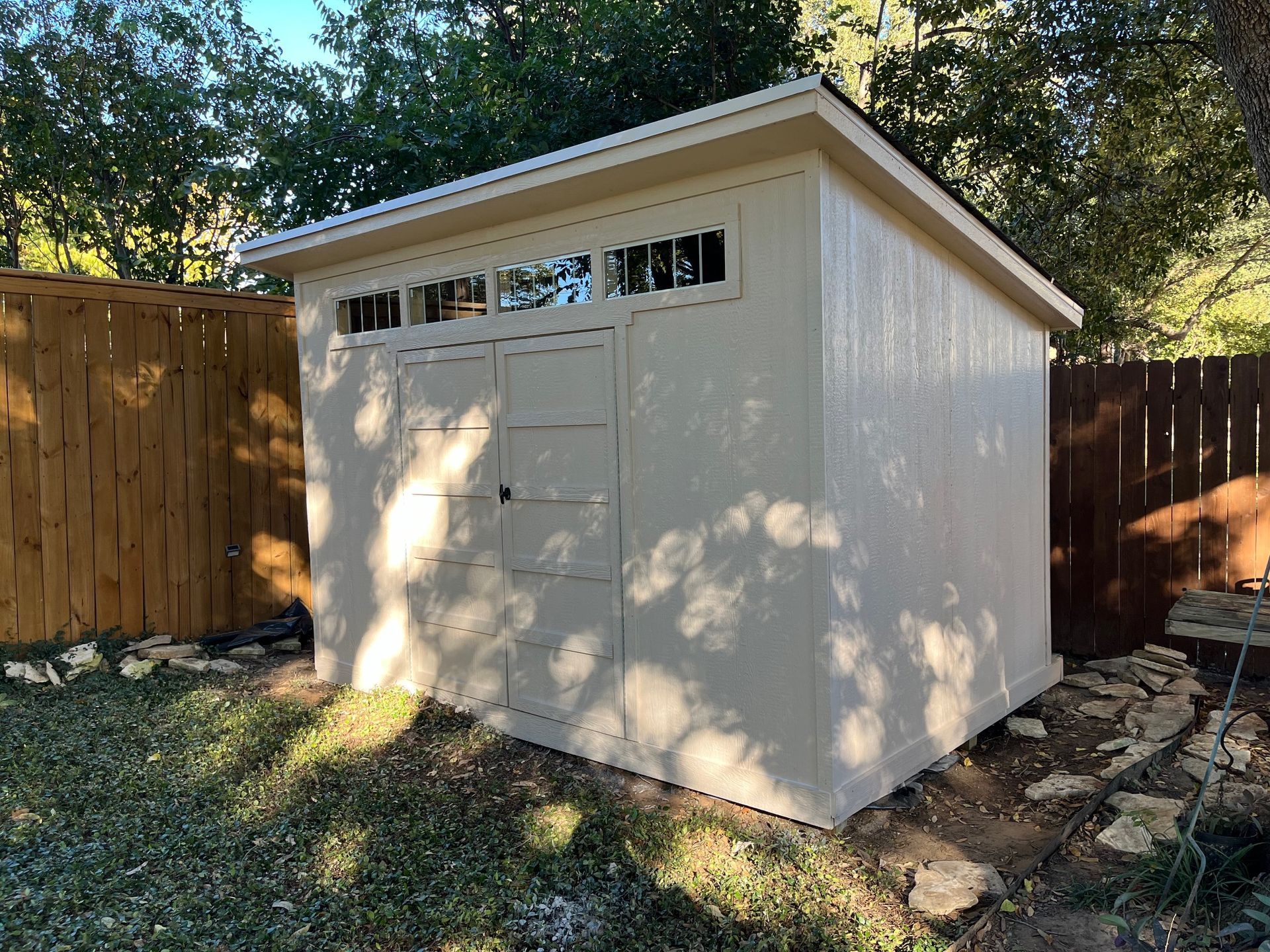 Tan shed with light-colored trim and a single window, set in a grassy yard, near a brown fence.