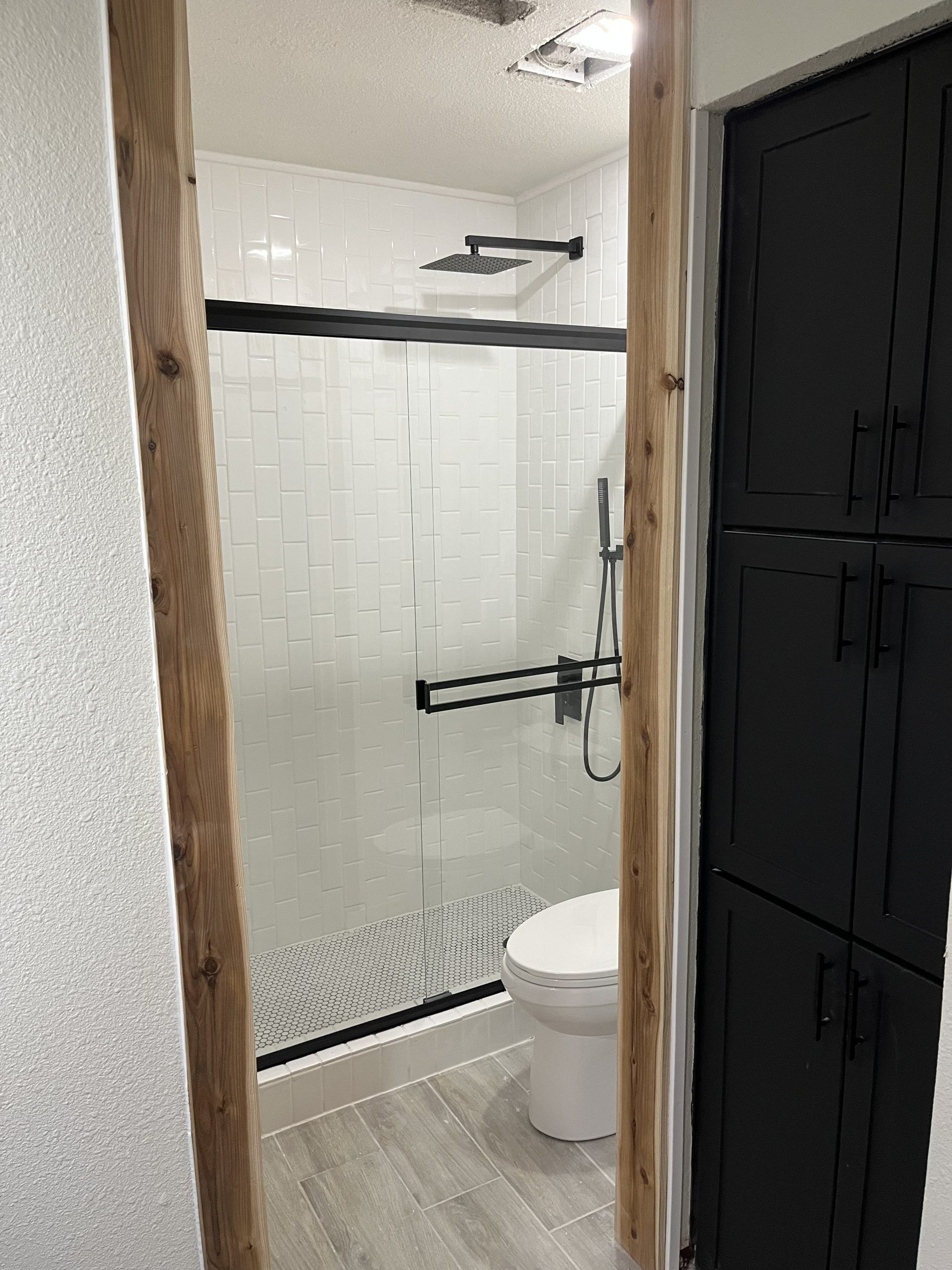 Bathroom with shower and toilet, black accents. Light-colored tile, wood trim, and black cabinets.