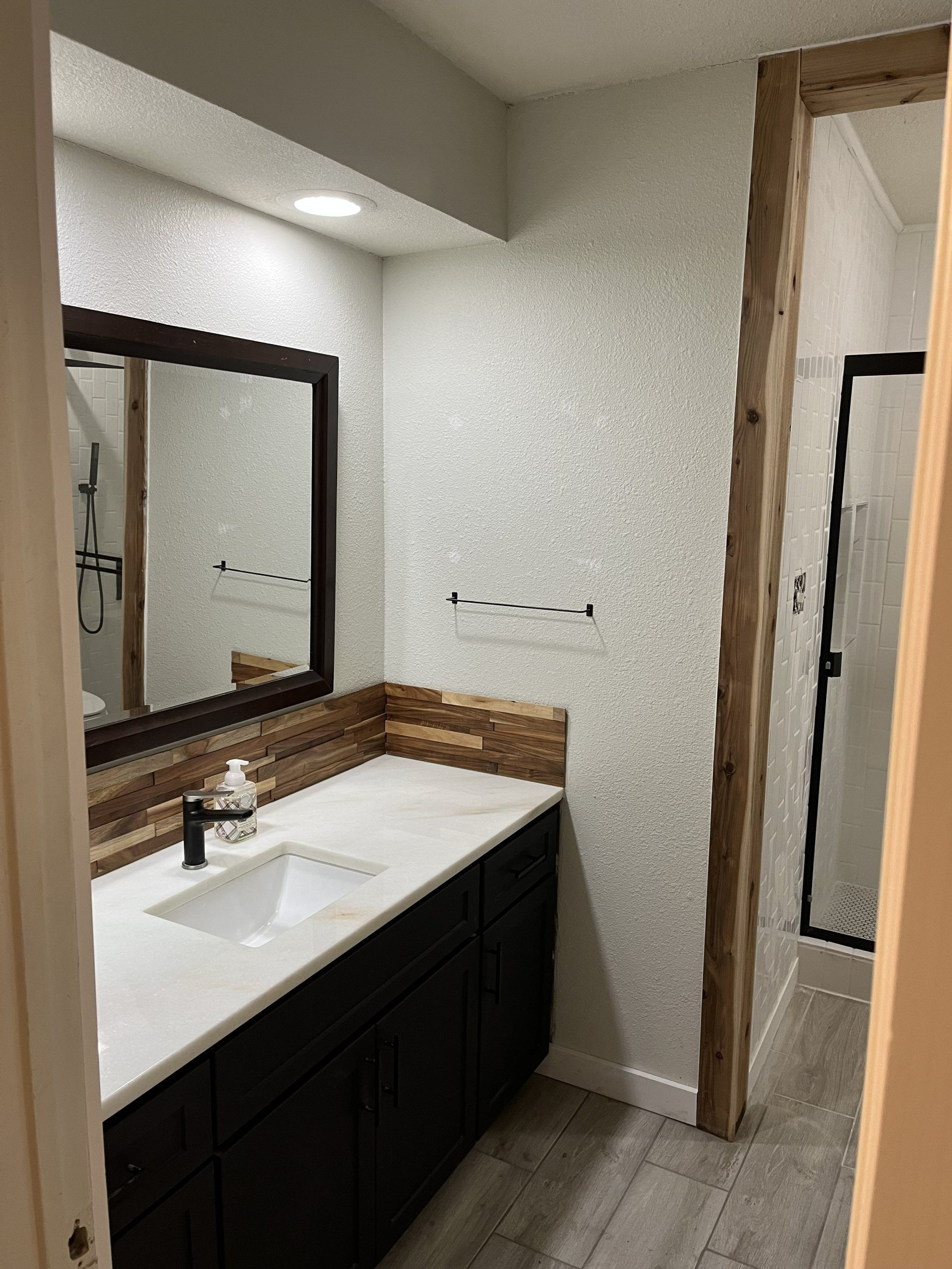 Bathroom with dark cabinets, white countertop, wood accents, and a view into a shower.