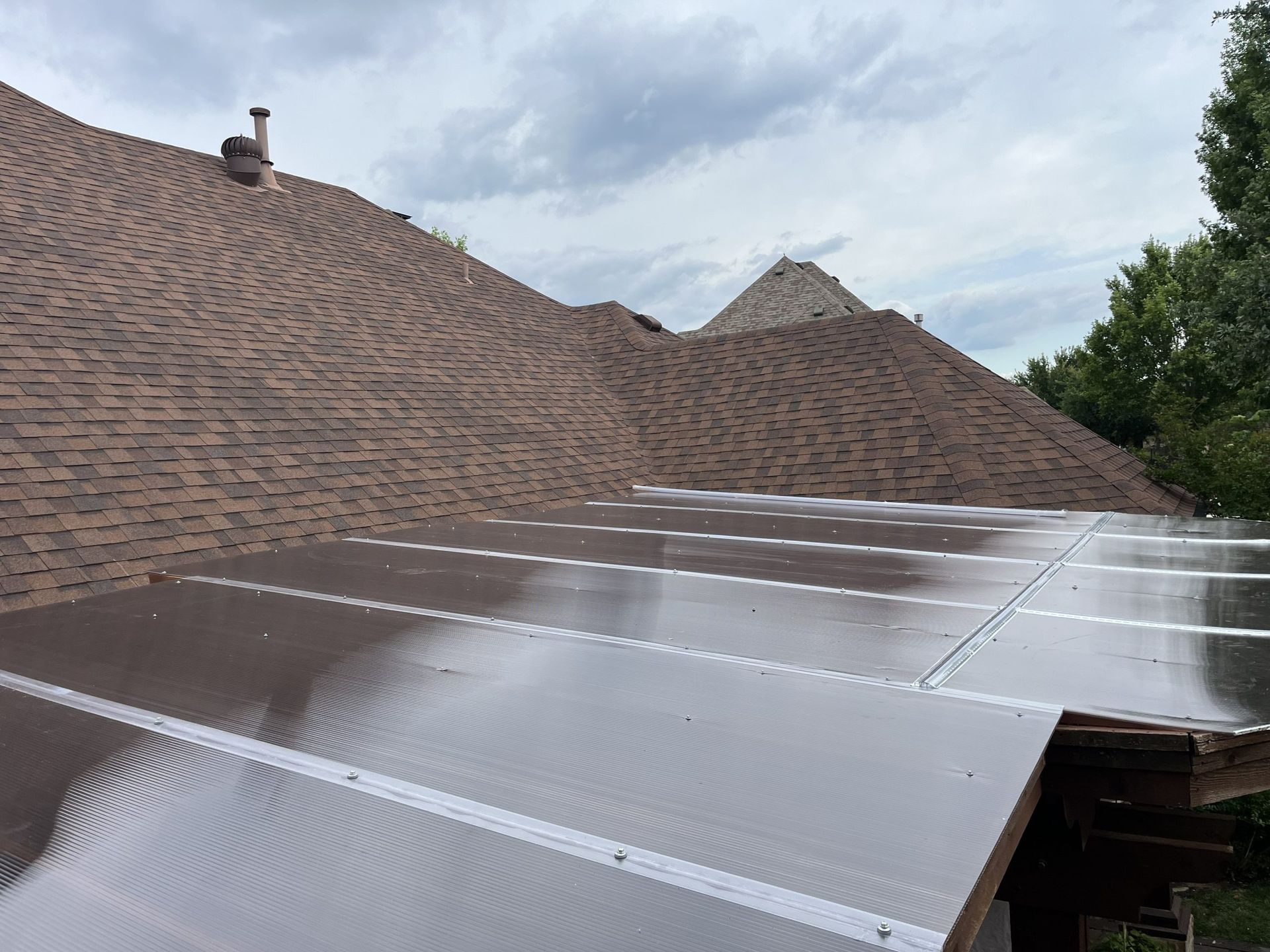 Brown shingled roof and transparent corrugated panels on a lower roof under a cloudy sky.