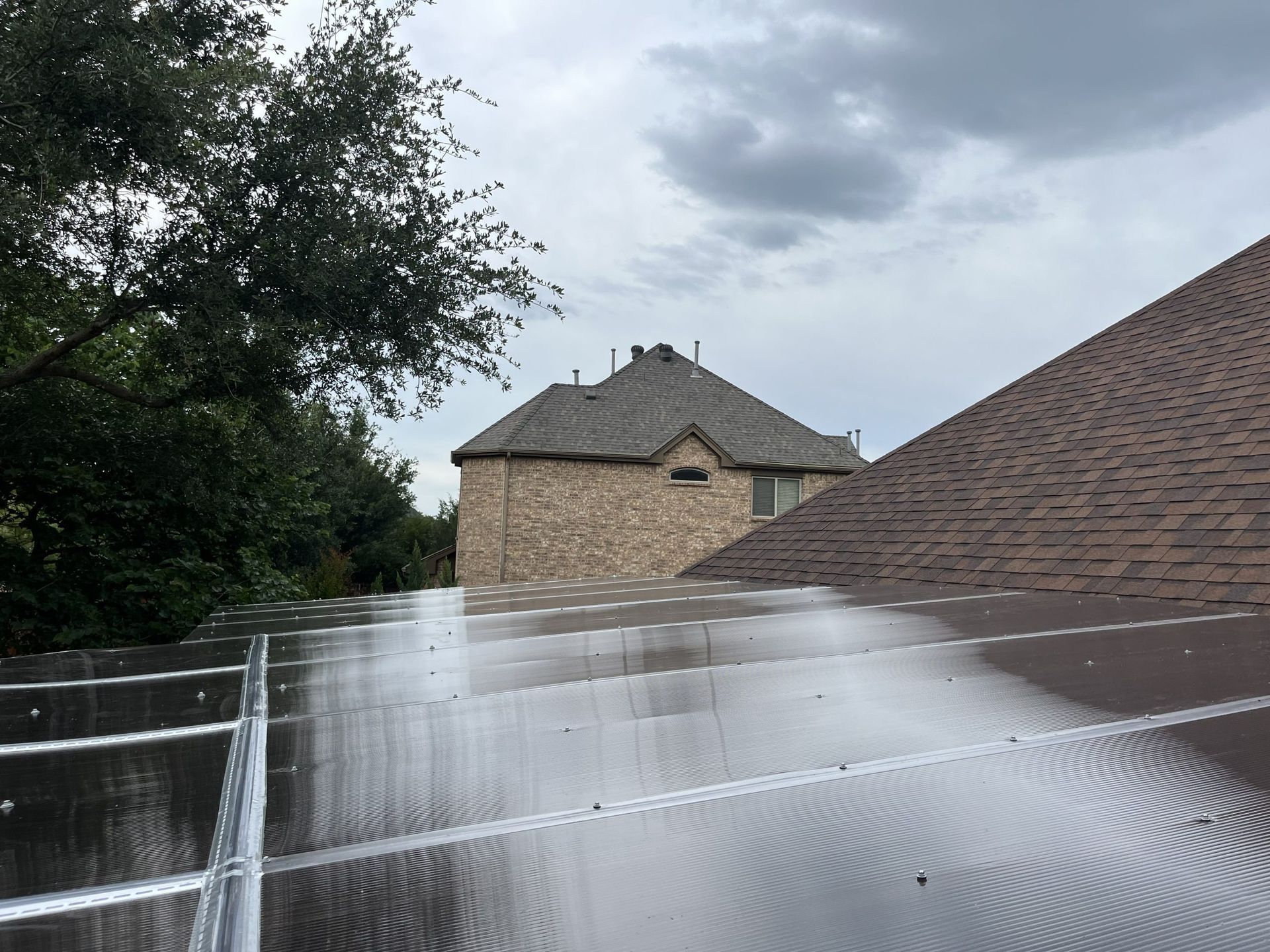 Rain-covered solar panels on a roof, with a brick house in the background under a cloudy sky.