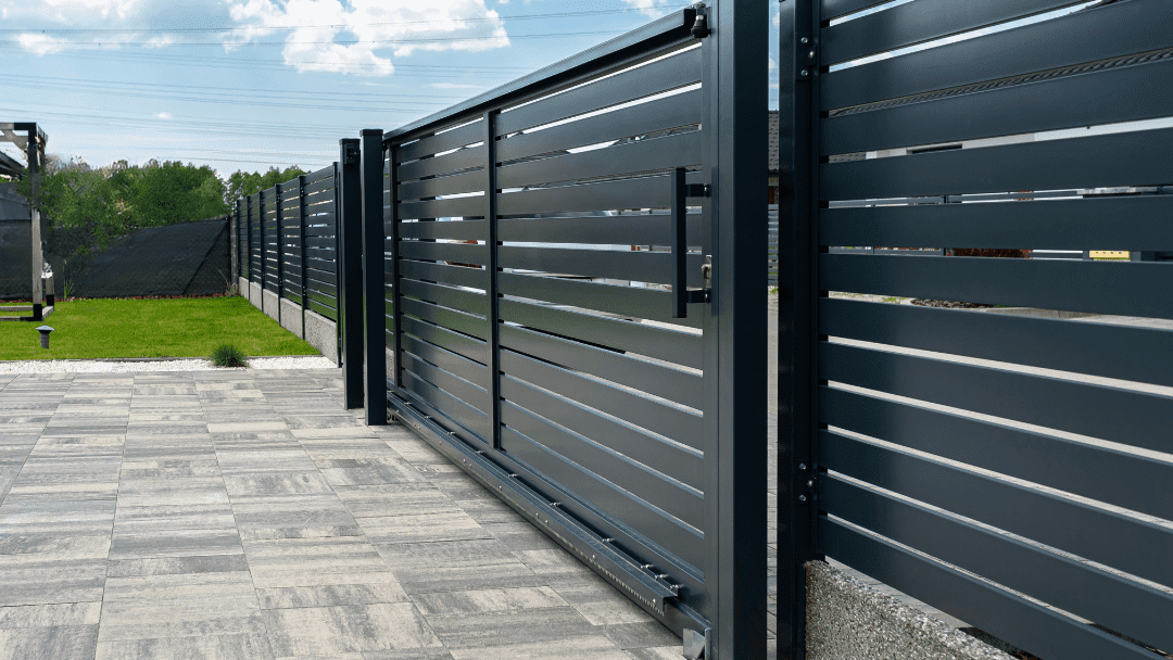 Dark gray horizontal slat sliding gate and fence on a brick driveway, under a blue sky.