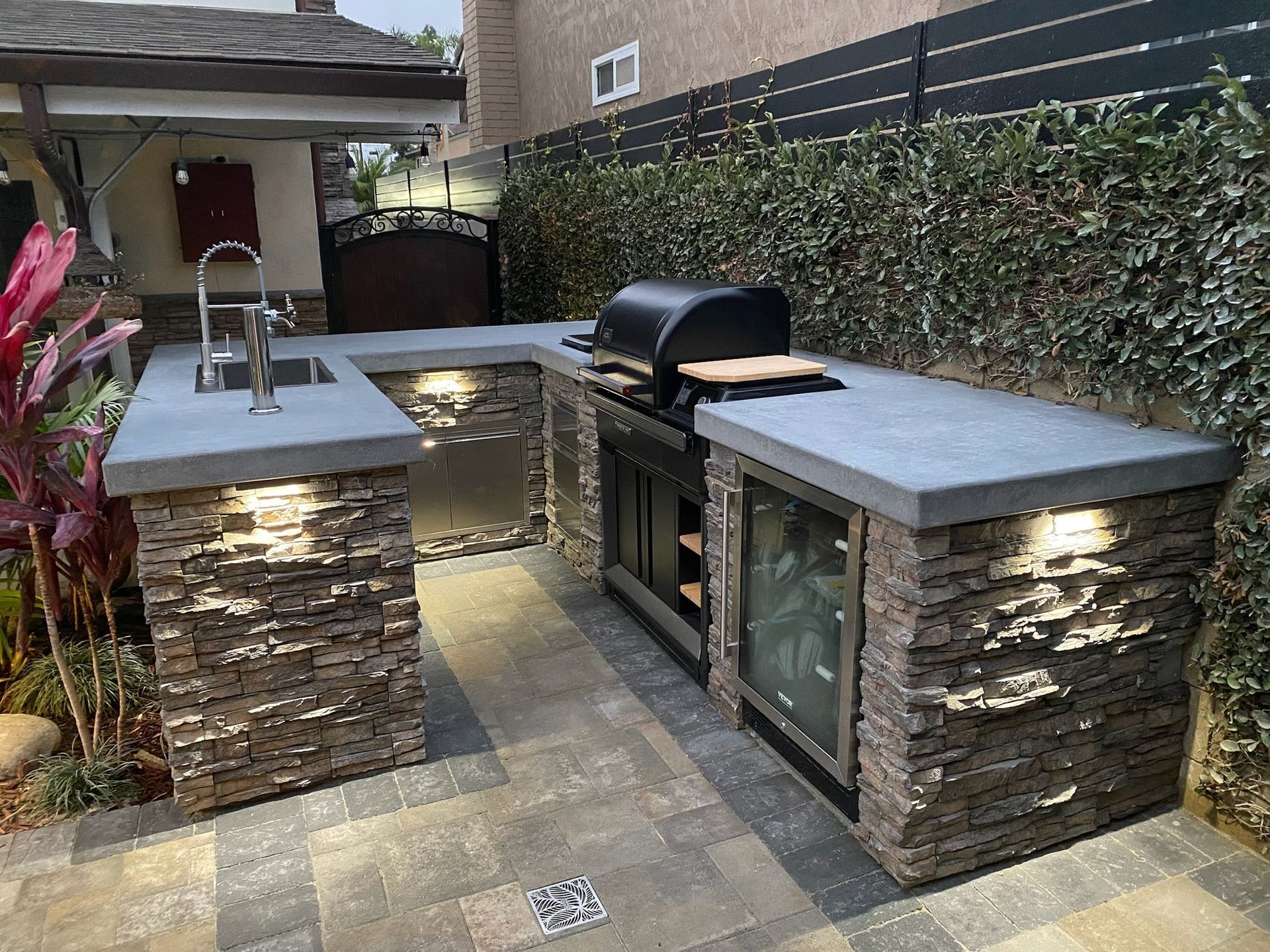 Outdoor kitchen with stacked stone, gray countertops, grill, sink, and beverage fridge, near a green hedge.