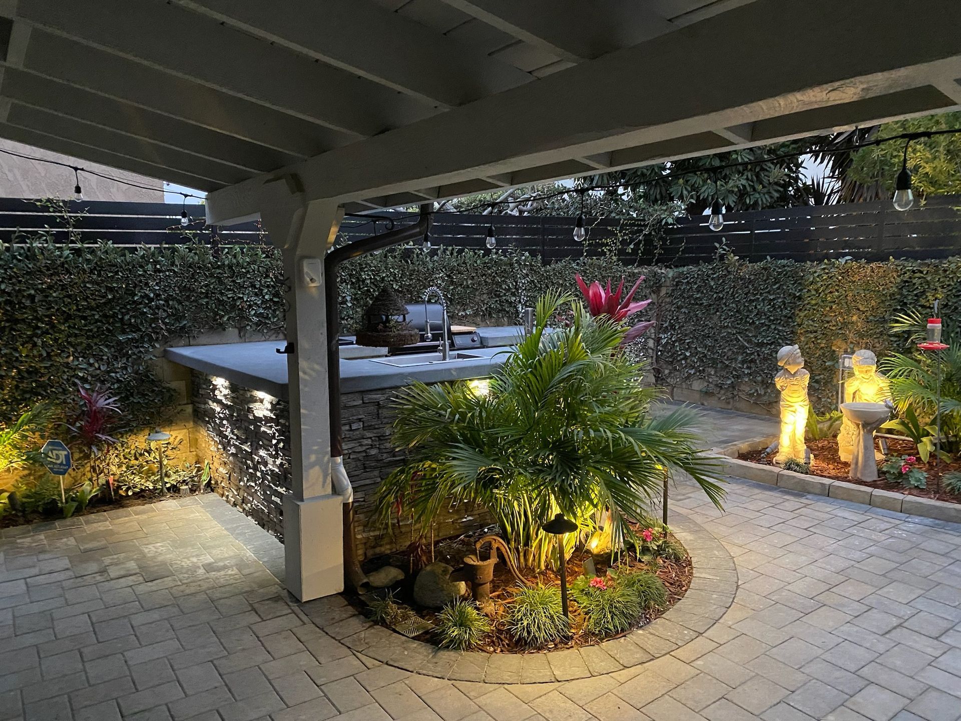 Nighttime backyard patio with lighting around a circular garden, bar, and statues; plants and hedges in view.