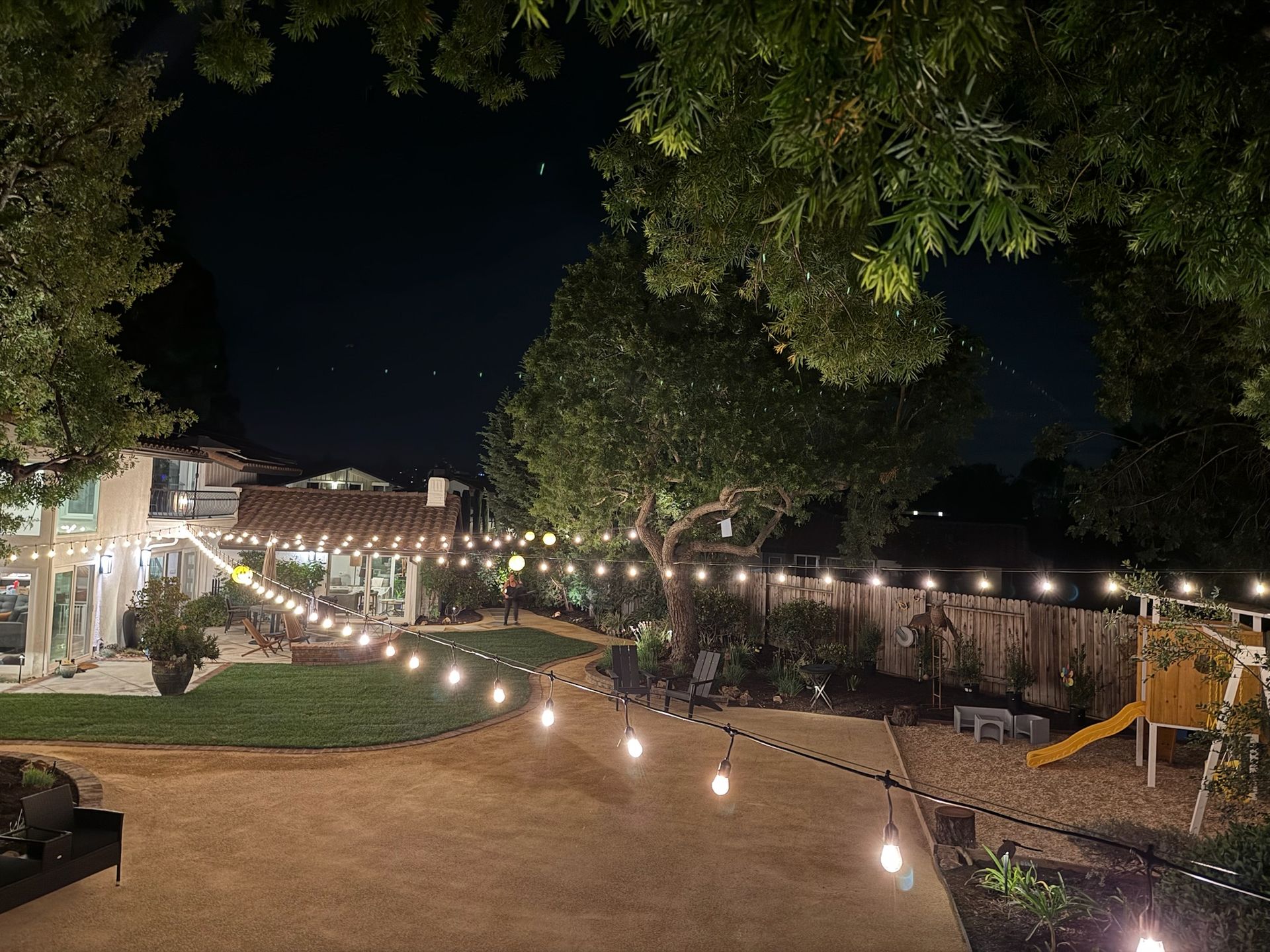 Night backyard with string lights, trees, lawn, play area, and house.