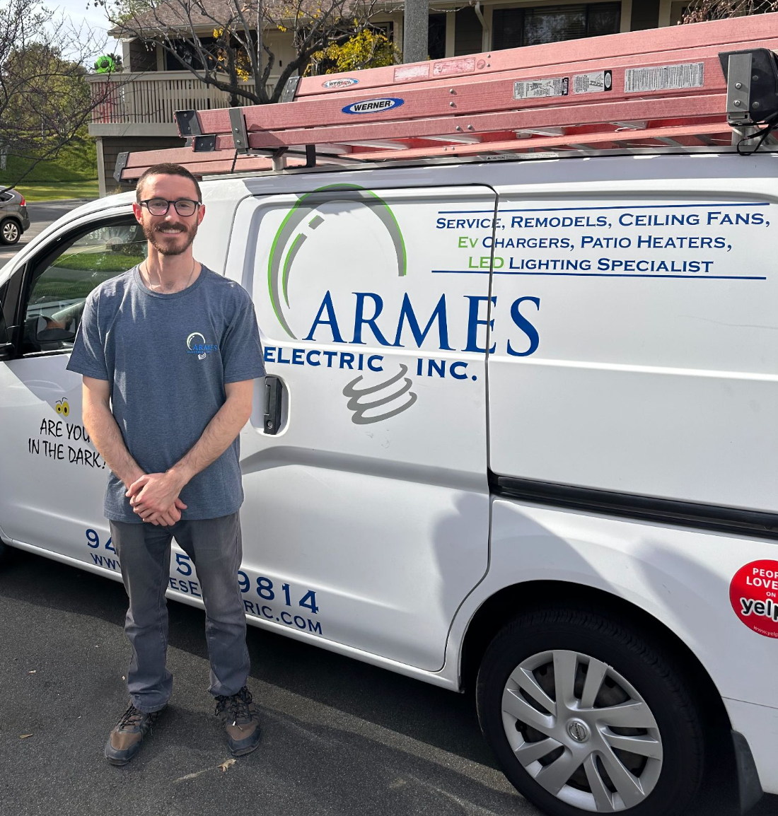 Man standing beside a white ARMS Electric van with a ladder on top, parked on a street.