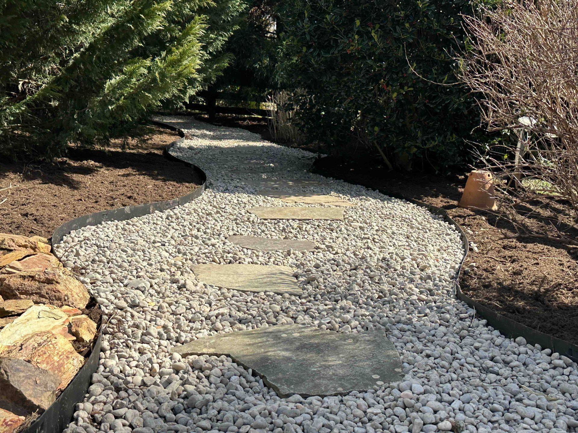 A winding garden path of white gravel with stepping stones, edged by dark borders and surrounded by trees and mulch.
