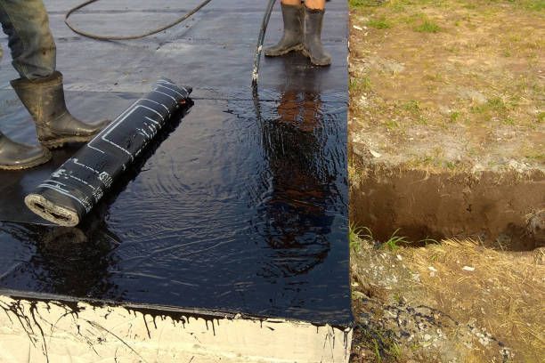 Workers applying black sealant to a concrete roof, next to an excavated area.