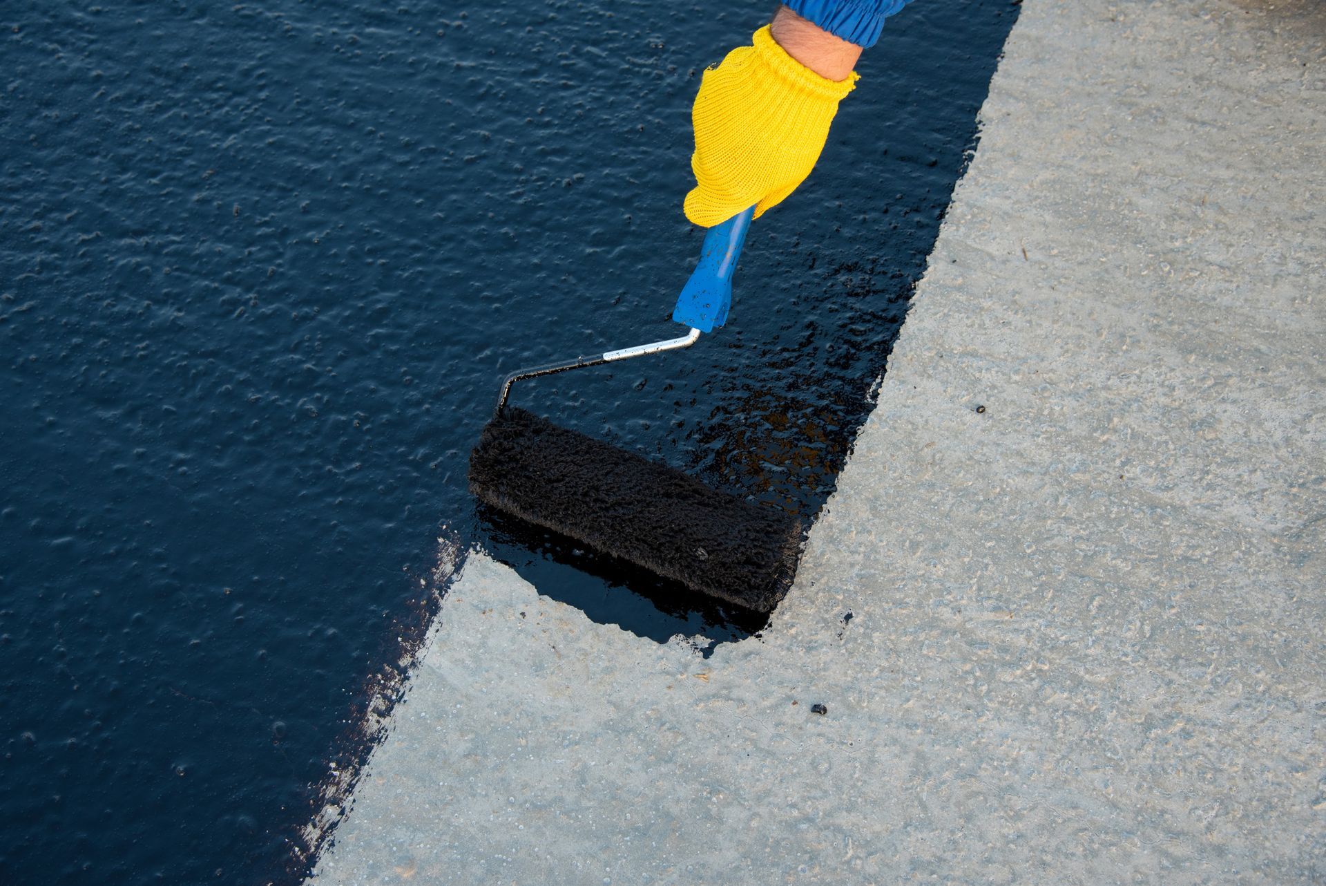 Person in yellow gloves rolling black sealant on a gray surface.