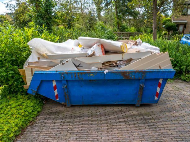 Blue dumpster filled with debris on a brick driveway, surrounded by greenery.