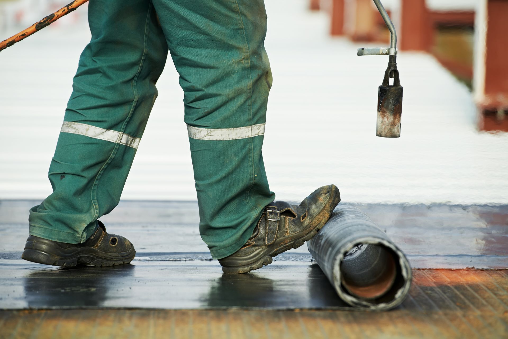 A worker in green overalls uses a torch to apply a black surface, likely roofing, near a metal tube.