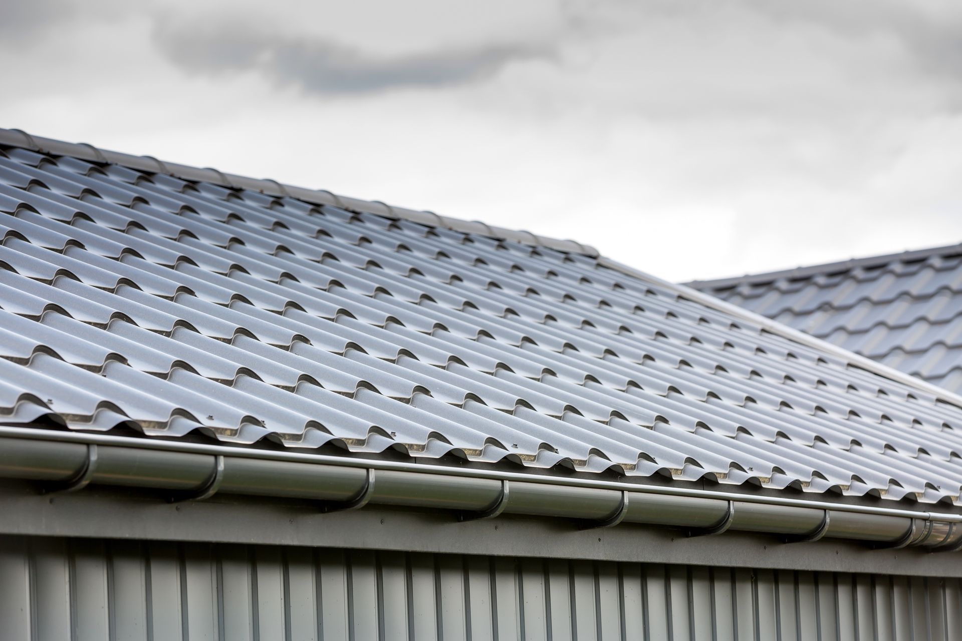 Gray metal tile roof with gutter and cloudy sky.