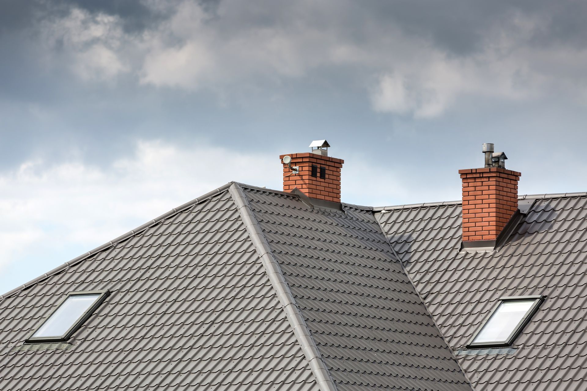 Gray tiled roof with two brick chimneys and two skylights against a cloudy sky.