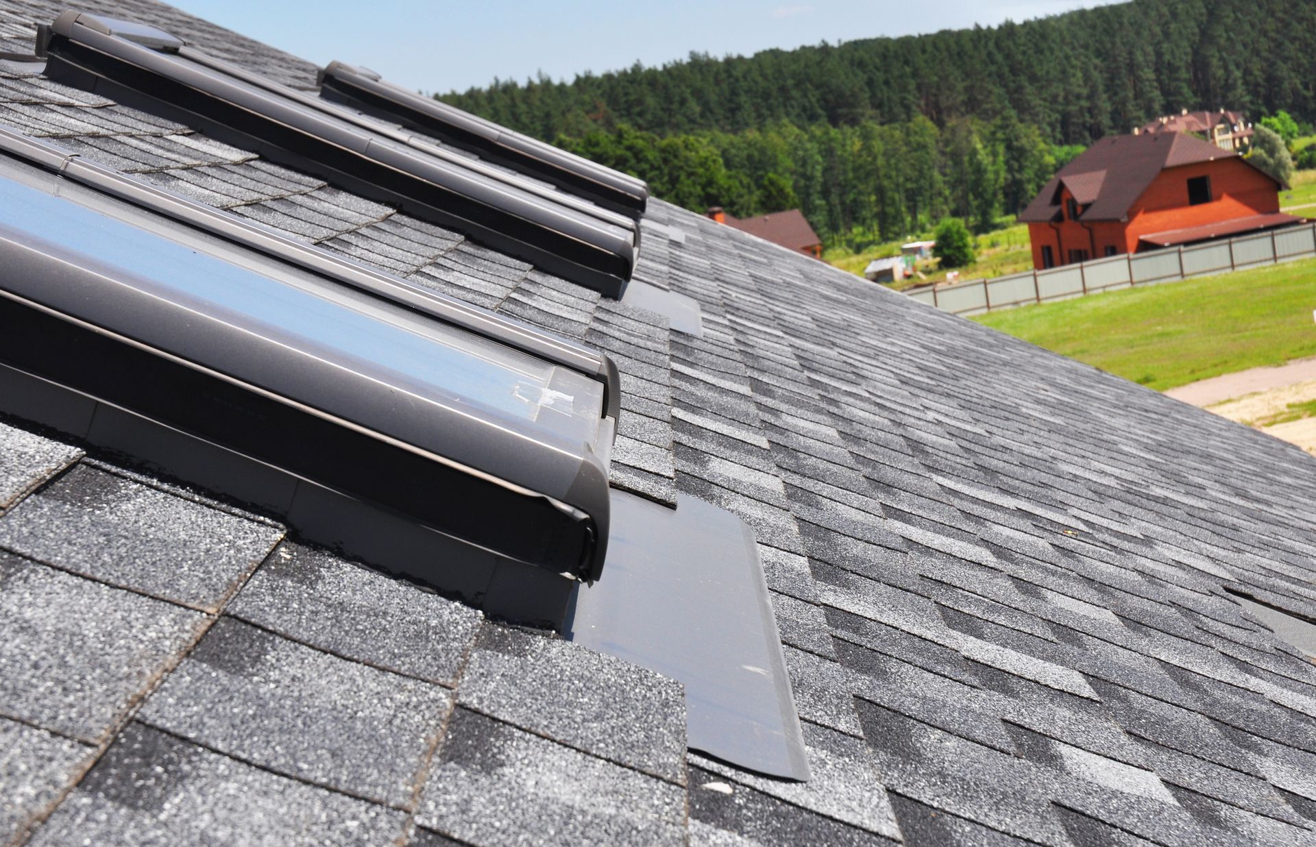 Gray shingled roof with several dark skylights; a house and trees are visible in the background.