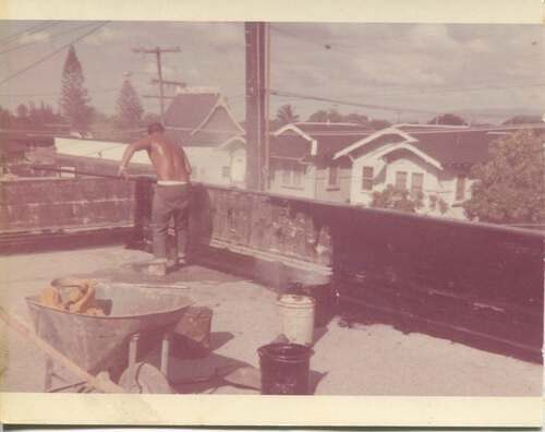 A shirtless man is standing on a balcony next to a wheelbarrow.