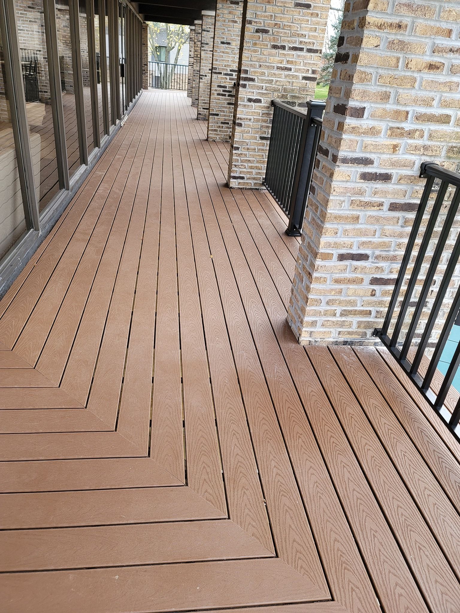 Brown composite deck with herringbone pattern, between brick columns and a railing.