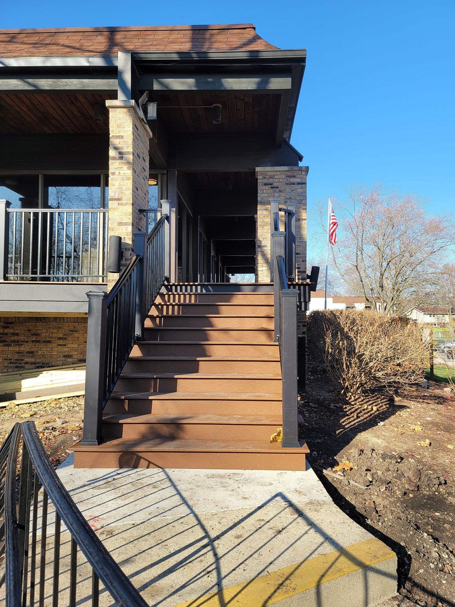 Outdoor staircase leading up to a building with a covered walkway. Sunny day, brown stairs, black railings.