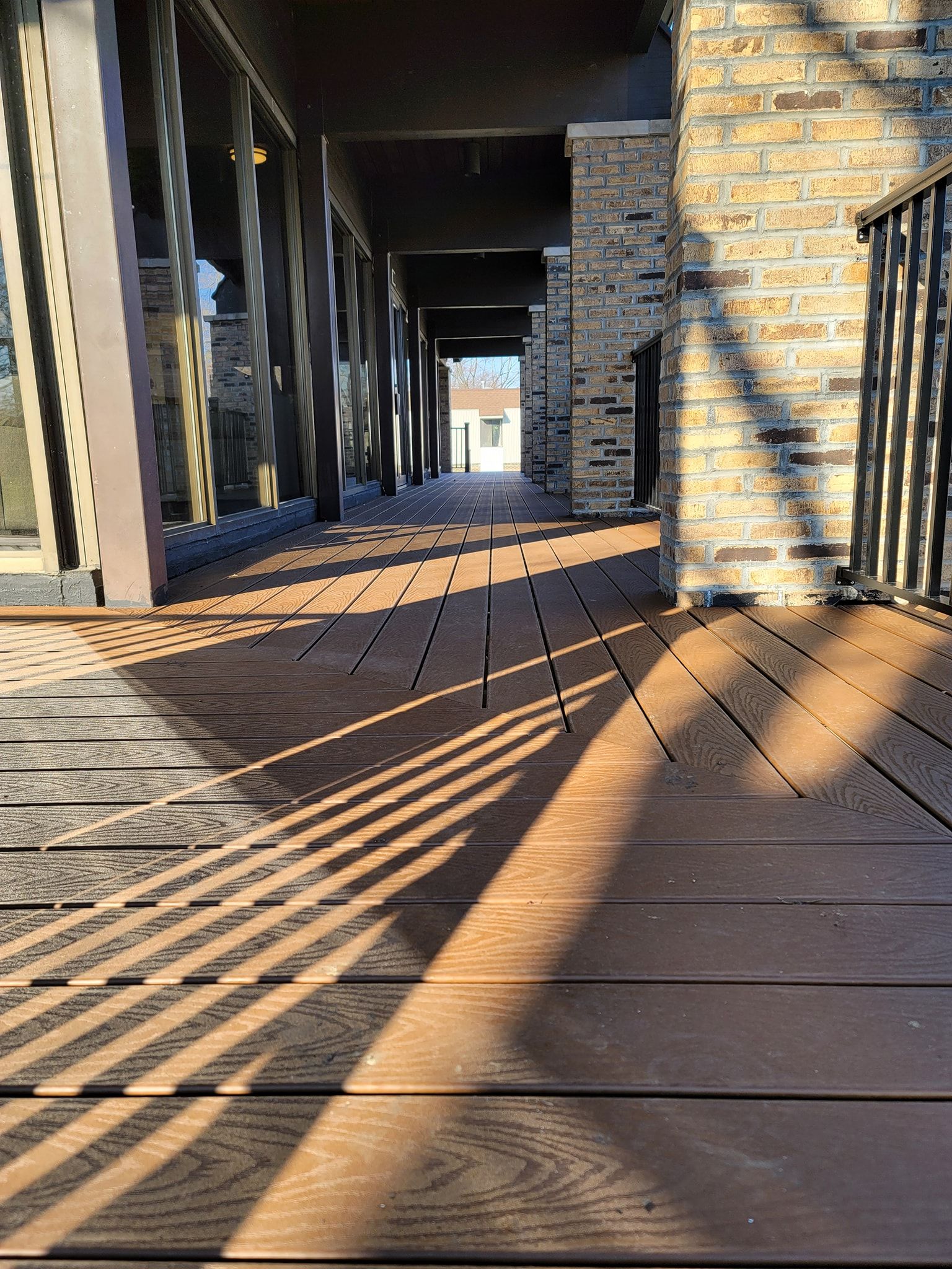 Wooden deck with shadows, partially covered by a structure with stone columns and railing.