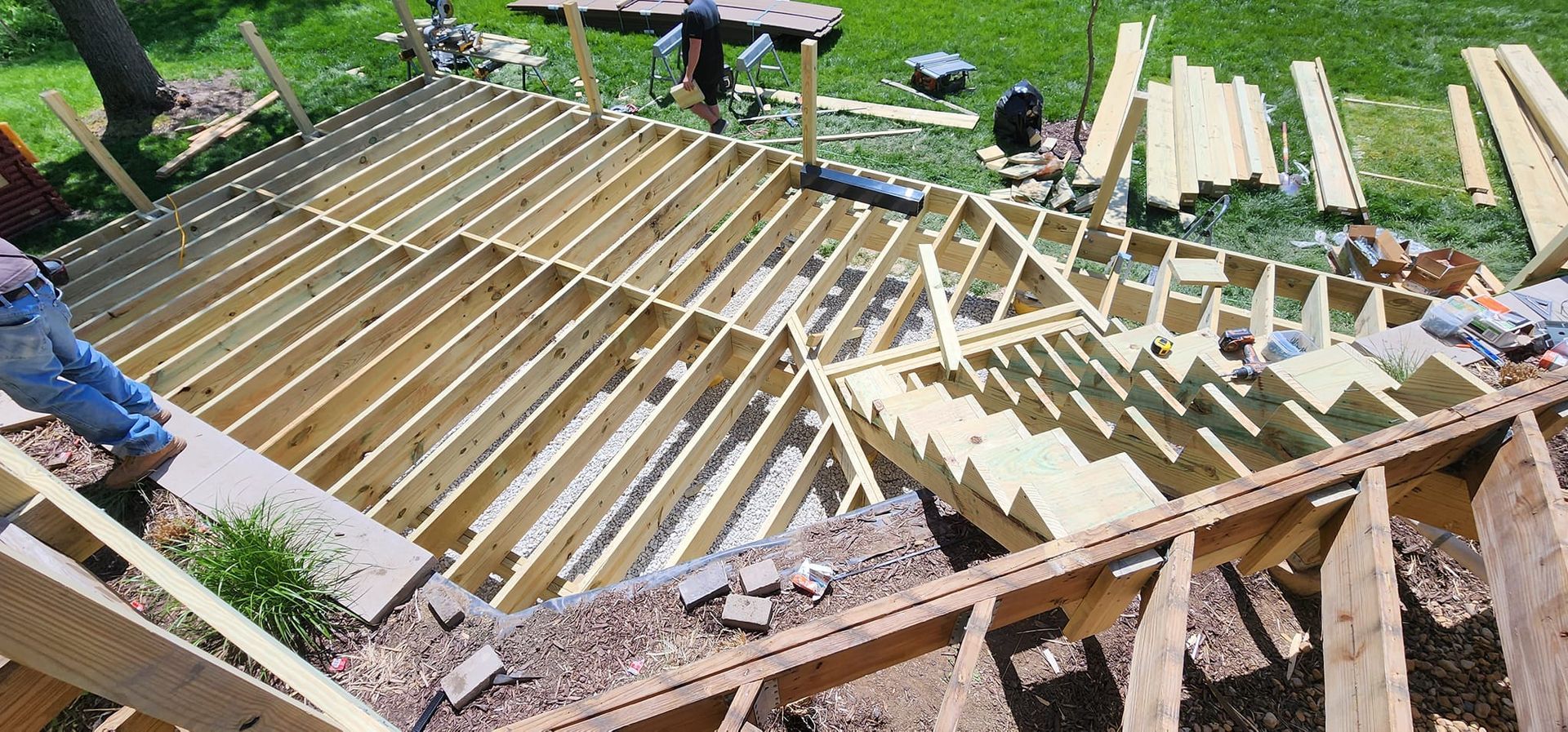 Construction of a wooden deck framework, with workers and materials on a grassy area.