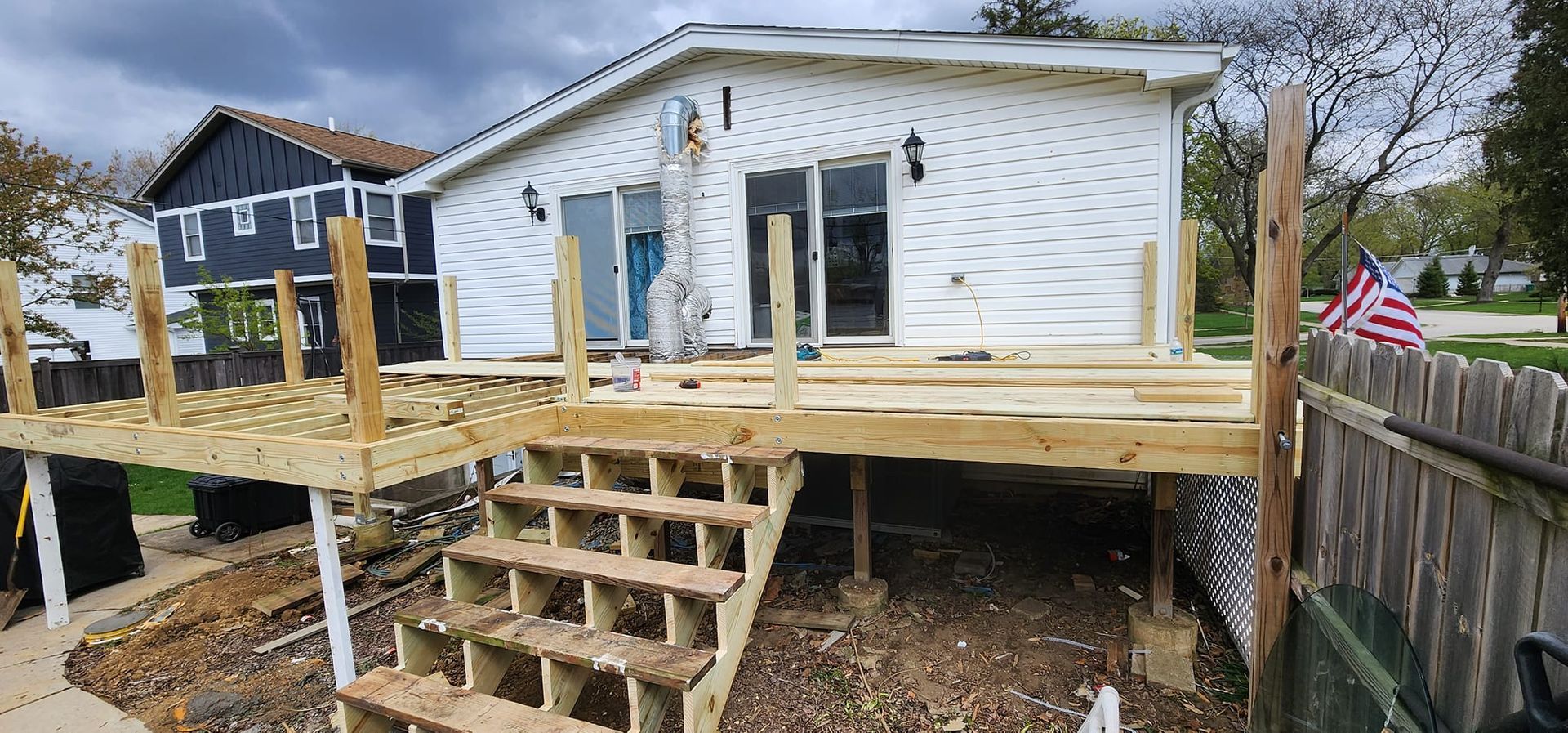 A partially built wooden deck with stairs in front of a white house. An American flag flies nearby.