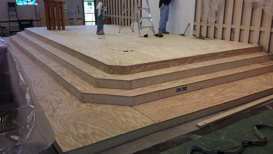 Wooden platform stage with three steps under construction; people working in the background.