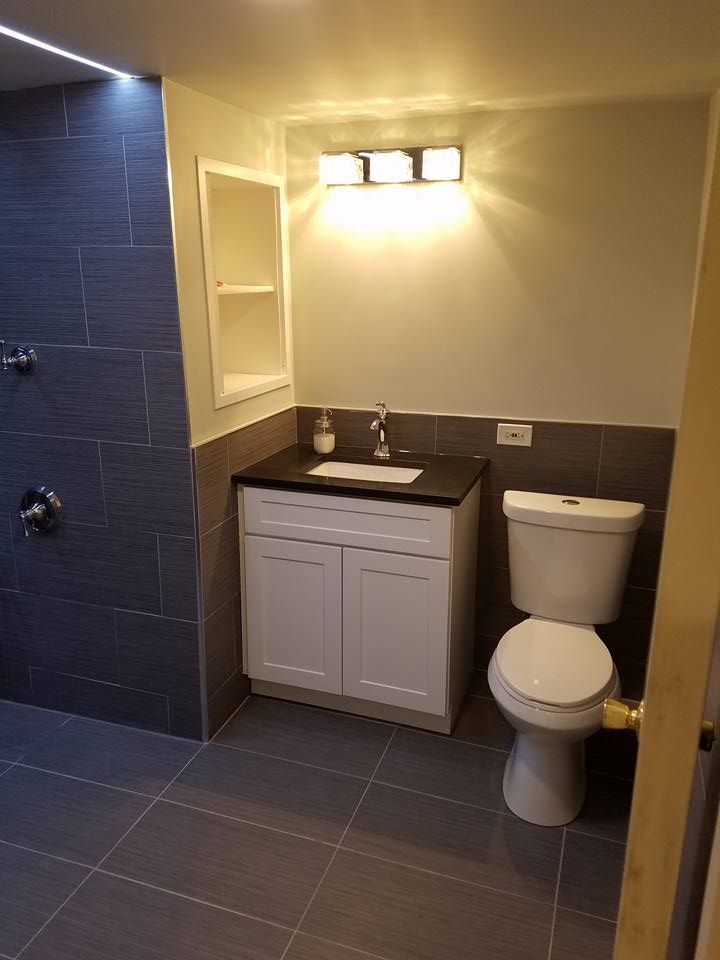 Bathroom with gray tile, white vanity, toilet, and shower fixtures. Lit by overhead lights.