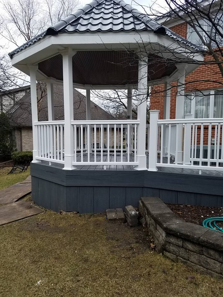 White gazebo with gray roof and deck, surrounded by grass and brick wall.