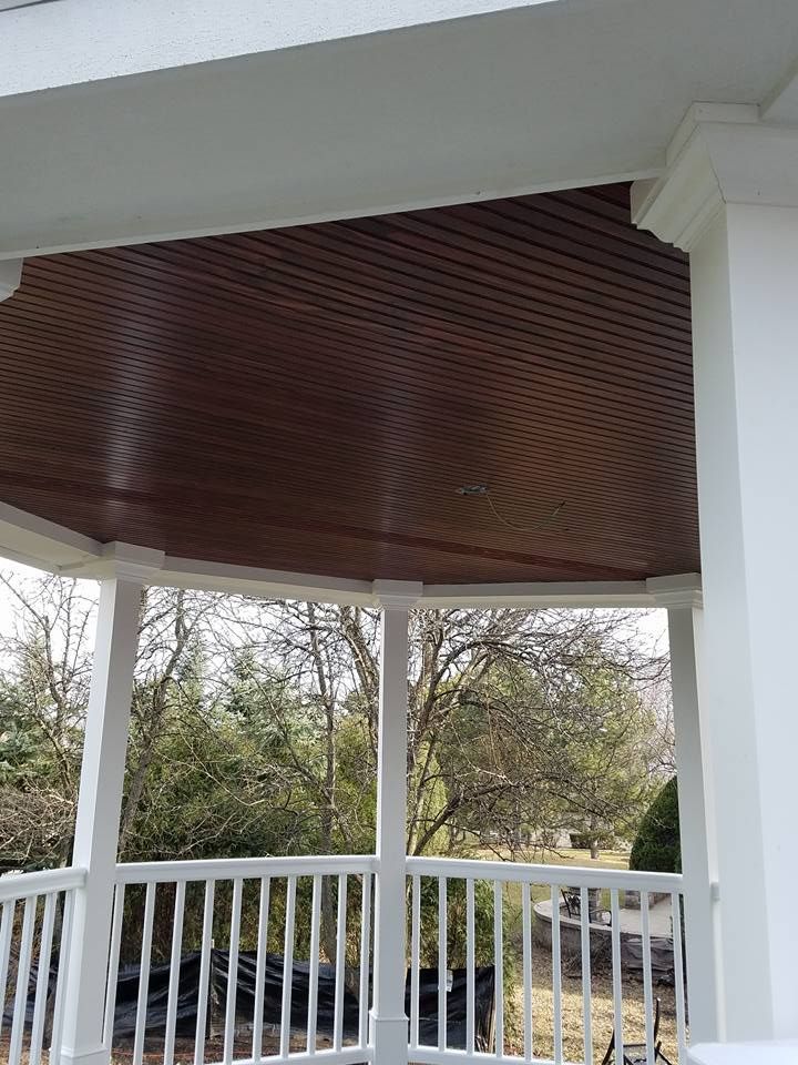 Brown wood ceiling of a porch, white columns and railing. Trees in the background.