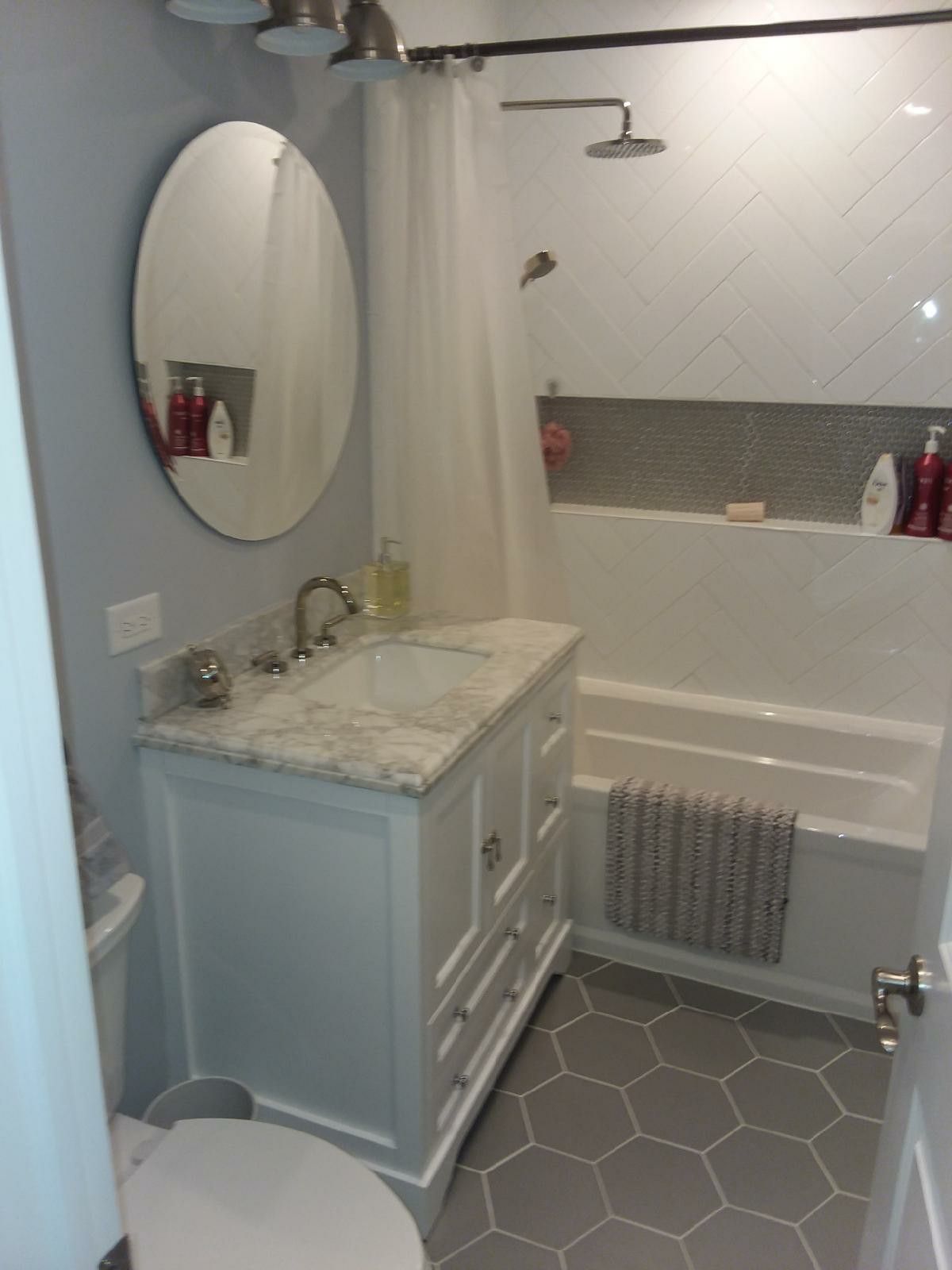 Bathroom with white vanity, oval mirror, white and gray tiled shower, gray hexagon floor tiles.
