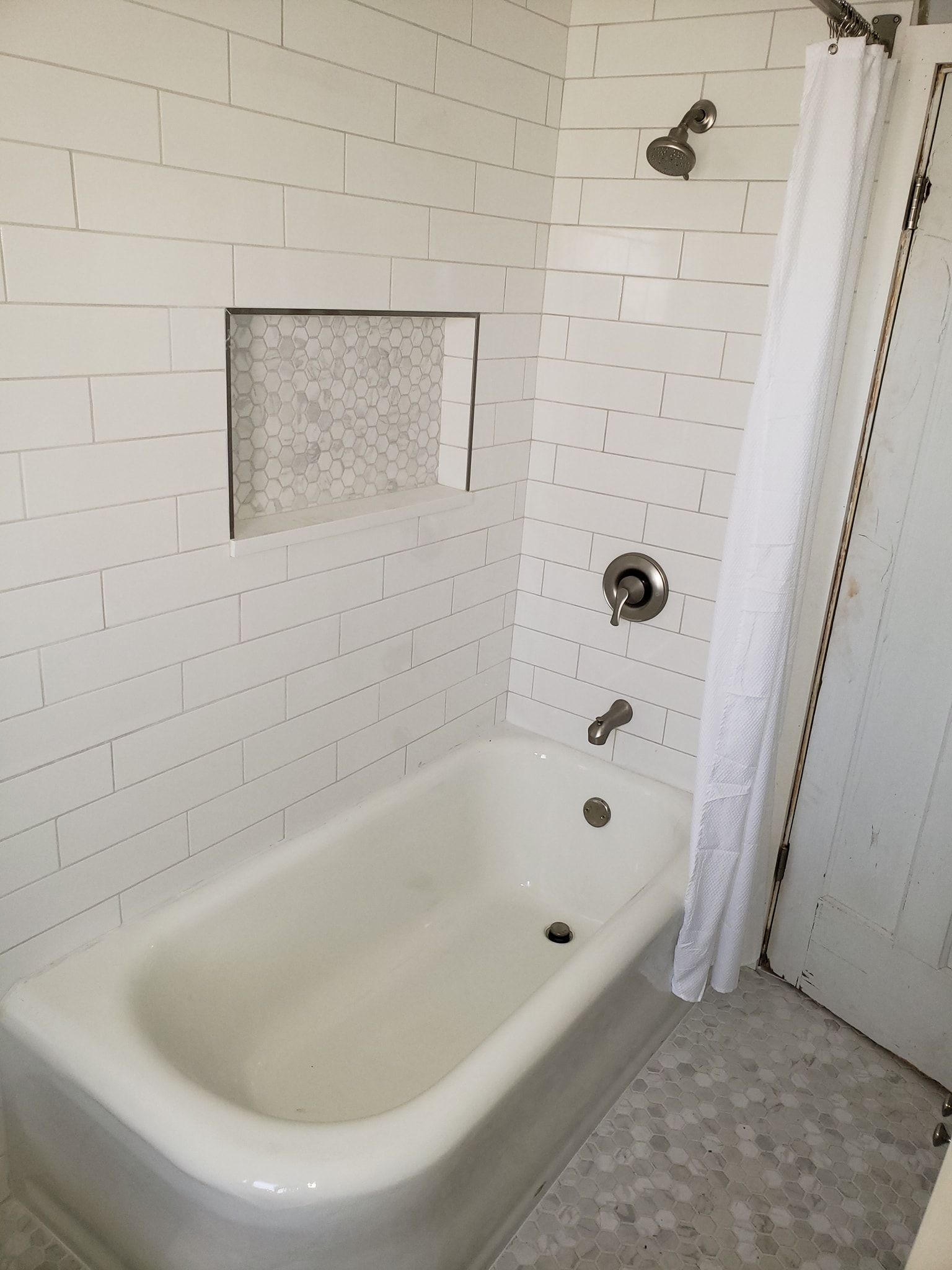 White tiled bathroom with tub and shower, built-in shelf, shower curtain, and silver fixtures.