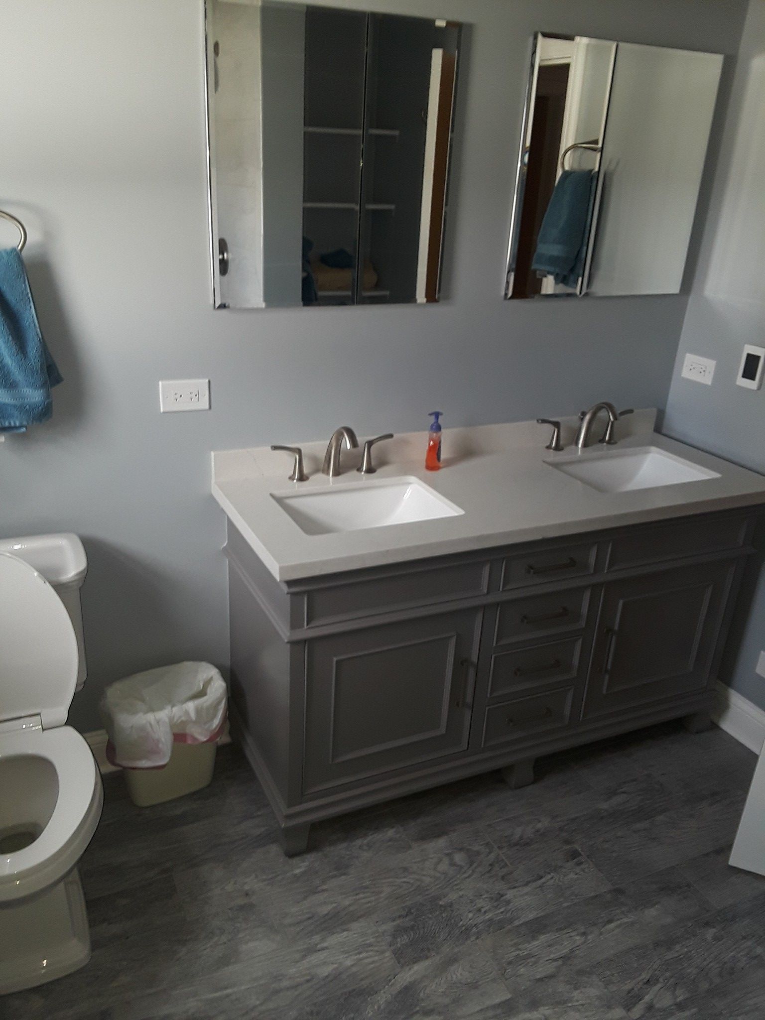 Bathroom with gray vanity, two sinks, and two mirrors on a gray wall. Toilet and wastebasket on the left.